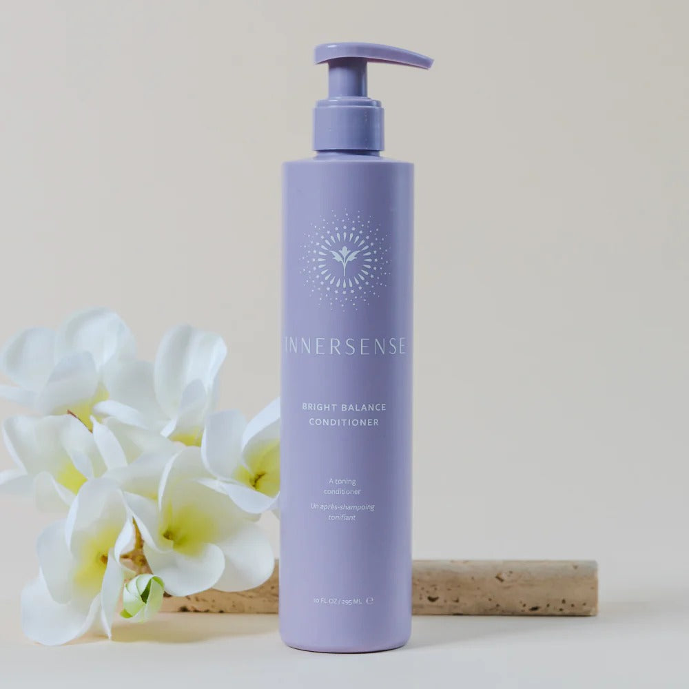 Innersense Bright Balance purple toning conditioner bottle with pump, next to white plumeria flowers.