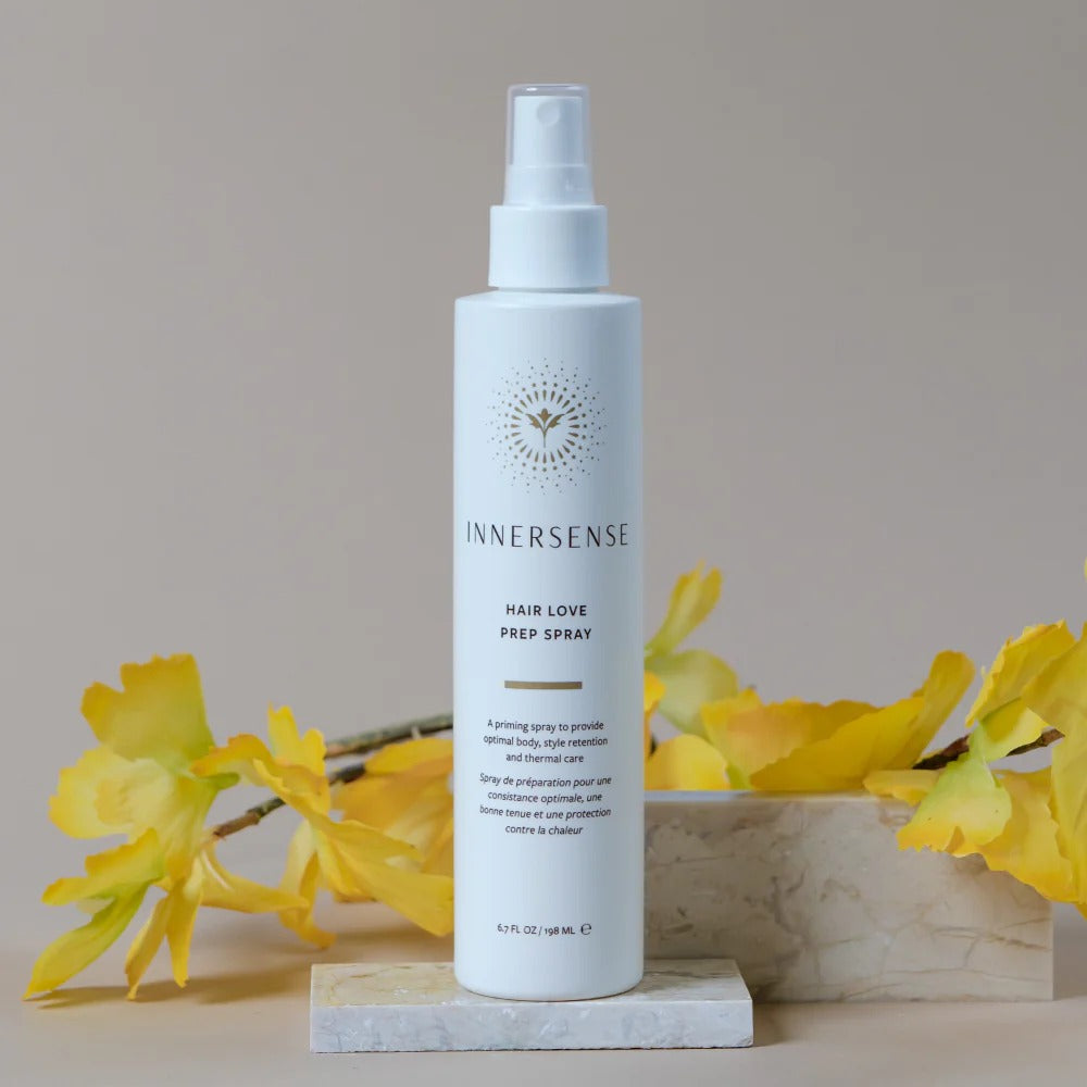 White Innersense Hair Love Prep Spray bottle with gold logo, surrounded by yellow flowers and marble blocks.