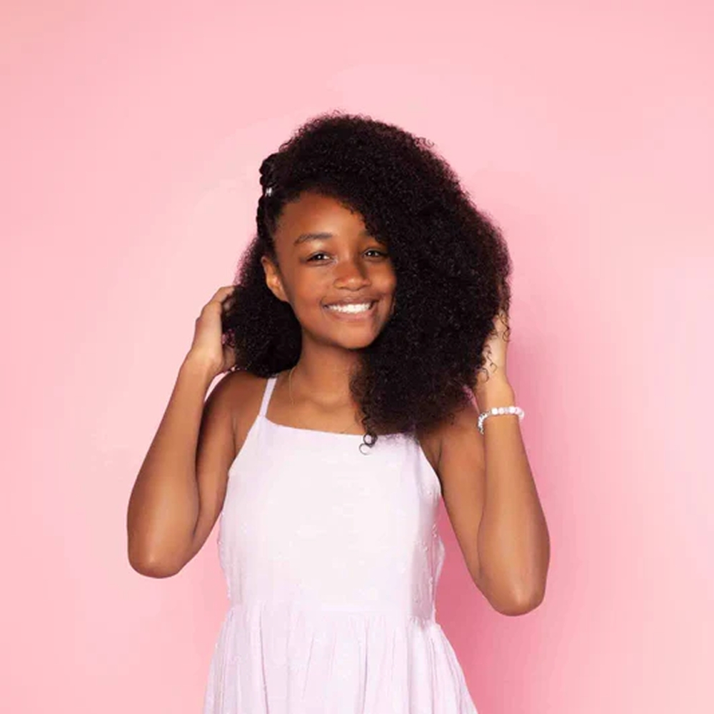 Smiling young girl with long, voluminous coily hair in a light pink dress against a pink background.