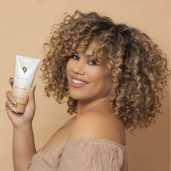 Smiling woman with curly hair holding a white and beige Bounce Curl leave-in conditioner tube.