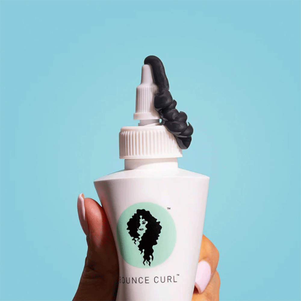 White Bounce Curl Turmeric Purifying Hair & Scalp Mask bottle with black product squeezing out, held against a blue background.