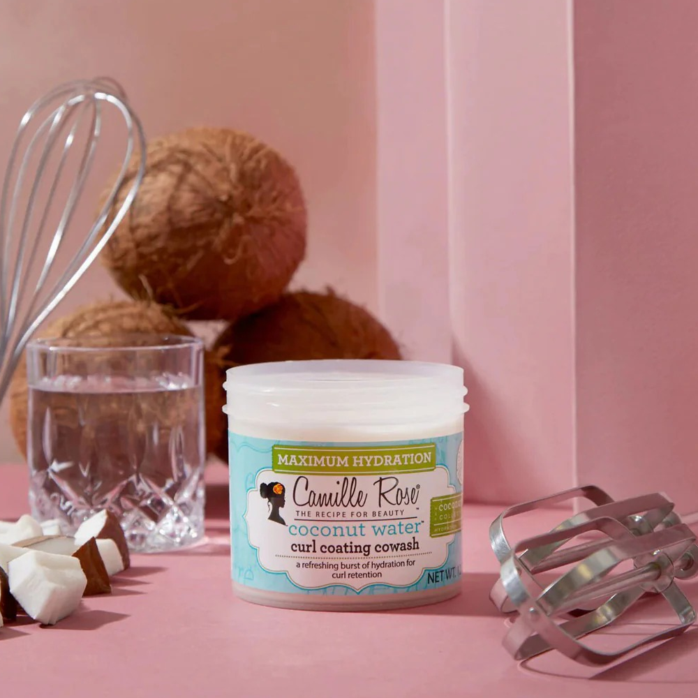 Camille Rose Coconut Water Curl Coating Co-Wash in a white jar, surrounded by coconuts, water, and whisks.